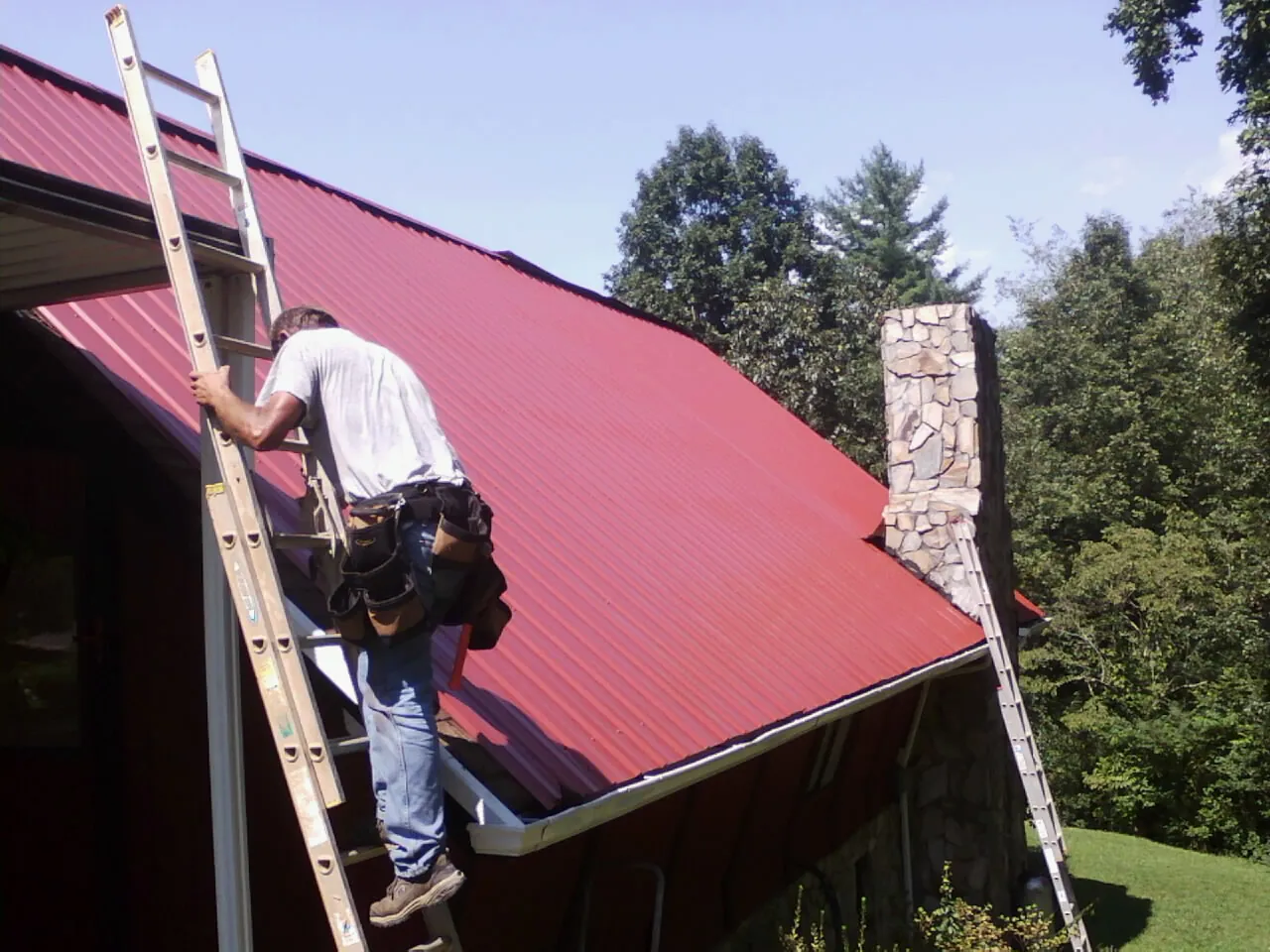 Roofer on a ladder beside a red metal roof and stone chimney — Soffit & Fascia Repair by Roofing Brothers in Parkers Prairie